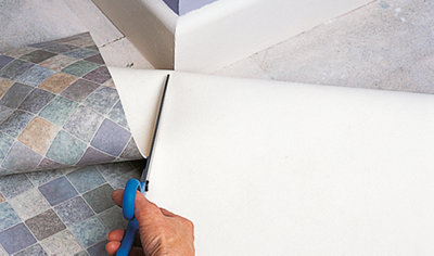 Sheet vinyl has been rolled back at the external corner of two walls. Vinyl is being cut in a straight line from the edge of the sheet to the corner of the two walls.