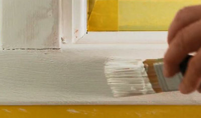 Close-up of a paintbrush being used to apply paint to a wooden window frame that has been sanded down.