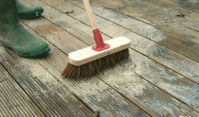 Person wearing wellies using a stiff broom to sweep the surface of a deck.