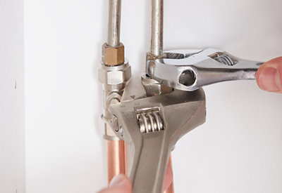A person using a spanner to loosen the nut where the kitchen tap tail joins the copper pipework, while a second spanner is used to steady the pipe.