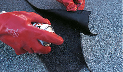Person wearing gloves lifting a piece of broken roof felt and spraying roof sealant underneath.