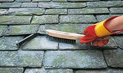 Person wearing gloves using a wire brush to clean the area around a broken slate roof tile.