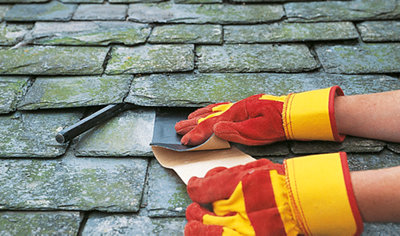 Person wearing gloves applying a piece of flashing strip to a slate tile and peeling off the backing paper. Tile above has been raised slightly using a bolster chisel.