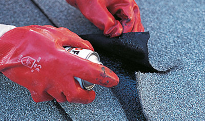 Person wearing gloves lifting a torn piece of roof felt and spraying roof sealant onto a new patch of roof felt that's been placed underneath.