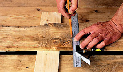 Person using a ruler and pencil to mark cutting lines on a floorboard that has been raised up with piece of wood slid underneath it.