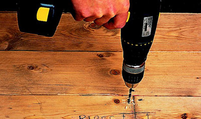 Person using a drill to drill a clearance hole close to the edge of a wood floorboard.