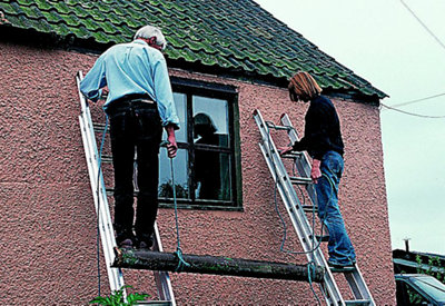Two people standing on ladders either side of a section of guttering. Guttering has been attached to a piece of rope at both ends, which is being use to lower the guttering to the ground.
