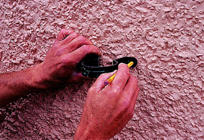 Person holding a gutter clip against a wall and marking the position of the fixing holes using a pencil.
