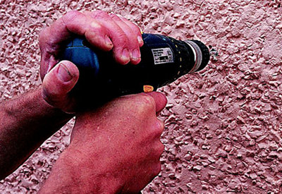 Person using a combi drill to drill a hole in the exterior wall of a house.