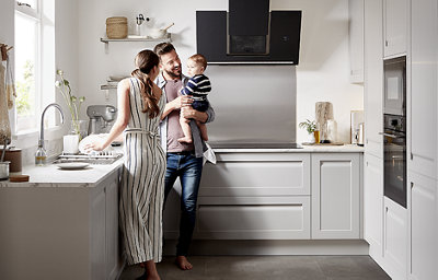 A woman is washing up at the kitchen sink while a man is holding a baby and looking happy. They are standing in a light grey U-shaped kitchen with white marble countertops.