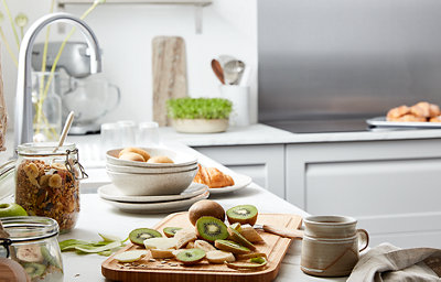 Two kiwi fruits have been sliced atop a wooden chopping board, placed on a white marble-effect countertop. Jarred granola and other plated foods surround the sliced kiwis. Behind, the chrome sink tap catches the light from the window.