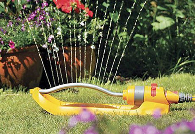 A sprinkler spraying water onto a lawn and the flowers and plants on the edge of the lawn.