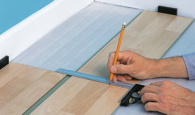Laminate board is pushed against the skirting board next to the laminate that has already been laid. Pencil and ruler are being used to mark where the board needs to be cut.