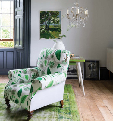 Light grey living room with an upholstered armchair with green leaf pattern, green rug, wooden flooring and countryside scene wall art.