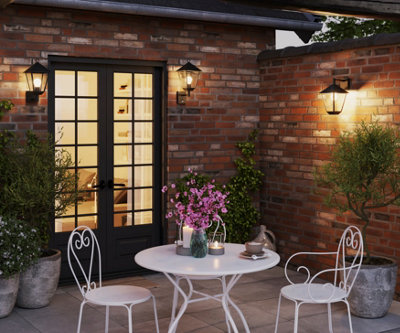 A small paved garden with a white 2 seater bistro set, and black wall mounted lights casting a warm glow throughout the garden.
