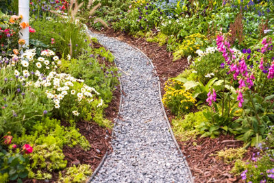 A narrow gravel pathway bordered by bark chips, surrounded by lush green shrubs and vibrant bedding plants.