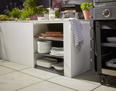 A rendered brick outdoor kitchen unit with a countertop and shelving underneath, holding plates and chopping boards. The outdoor kitchen unit is next to a barbecue.