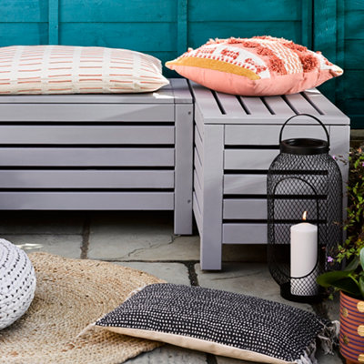 A grey-painted corner garden bench sits on a patio in front of a teal-painted fence, creating a cozy outdoor seating area.