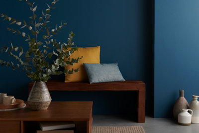 A coffee table with a large plant in a vase, a bench with two cushions on, in front of a wall painted a deep navy blue.