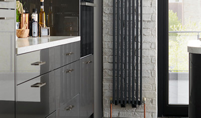 Black vertical radiator mounted on white brick wall in grey kitchen.