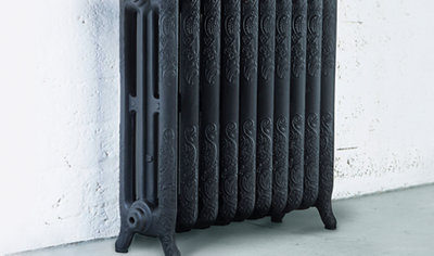 Black cast iron column radiator sitting on grey floor in front of white brick wall. Radiator has an intricate pattern on the front.