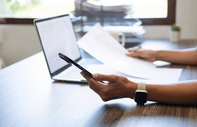 Person sat at a desk with a phone in one hand, peices of paper in the other hand and a laptop sat on the desk.