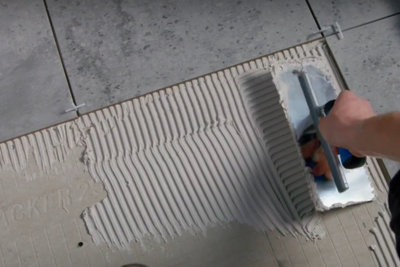 Person using a notched trowel to spread tile adhesive onto the top of tile backer boards. Notched trowel has created grooves in the adhesive.