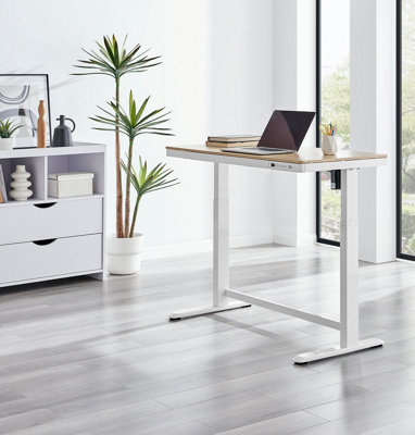 A white standing desk in a large home office with white walls, floor to ceiling windows and light grey wood effect laminate flooring.