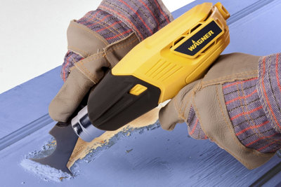 A person wearing gloves using a heat gun and scraper to remove paint. The heat gun is focused on an area where the paint has started to bubble, indicating it's loosening from the surface for easy removal.