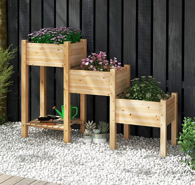 Wooden three-tiered planter filled with bedding plants, placed beside a black exterior wall.