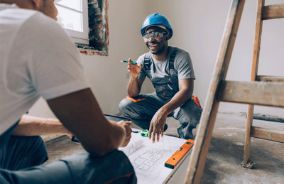 Tradesperson with hard hat kneeling talking to another person in a room which is being renovated